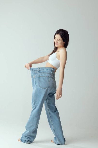 Full-length portrait of young slim woman wearing giant jeans, posing isolated over grey studio background. Weight-loss, figure concept - Fotografie, Obrázek