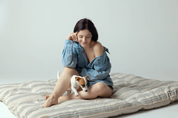 Portrait of young beautiful woman sitting on mattress, posing with cute small dog, puppy isolated over grey studio background - Fotoğraf, Görsel