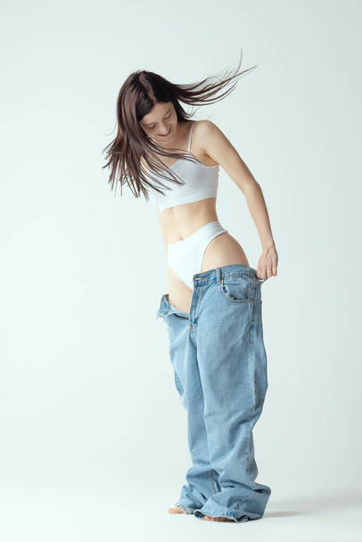 Full-length portrait of young slim woman wearing big jeans, posing isolated over grey studio background. Weight-loss concept - Фото, зображення