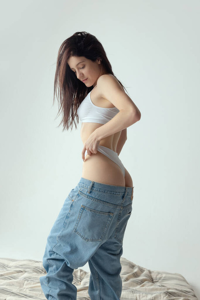 Portrait of young beautiful woman posing in underwear and jeans isolated over grey studio background. Perfect body - Foto, immagini