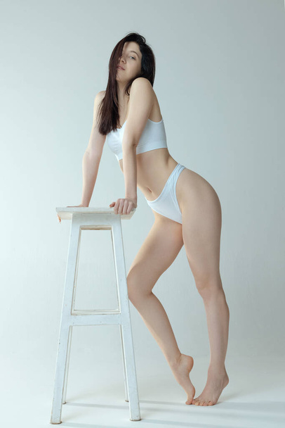 Dreamy looking girl leaning on chair, posing in white underwear isolated over grey studio background. Tendernes - Fotó, kép