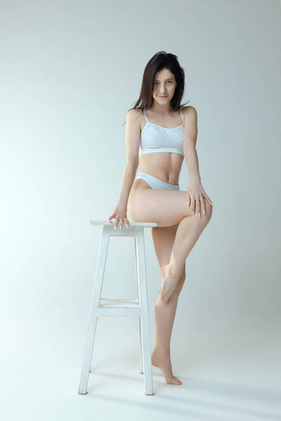 One tender young girl sitting on chair in white cotton underwear, posing isolated over grey studio background - Foto, Imagen