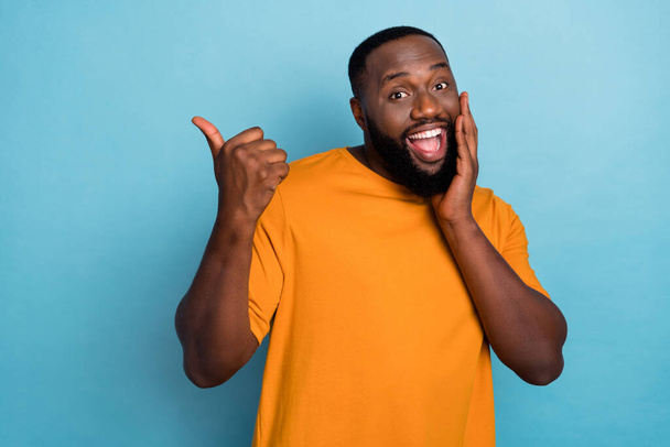 Photo of cool impressed guy wear orange t-shirt pointing thumb up empty space arm cheek isolated blue color background - Fotó, kép