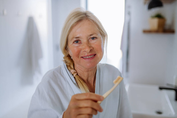 Beautiful senior woman in bathrobe brushing teeth with eco wooden toothbrush inbathroom, sustainable lifestyle. - Foto, afbeelding
