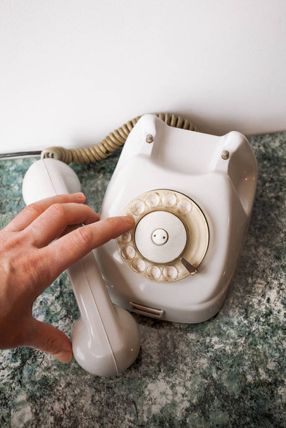 Un dito compone un numero su un vecchio telefono sovietico bianco analogico su un piano d'appoggio strutturato blu e uno sfondo bianco. - Foto, immagini