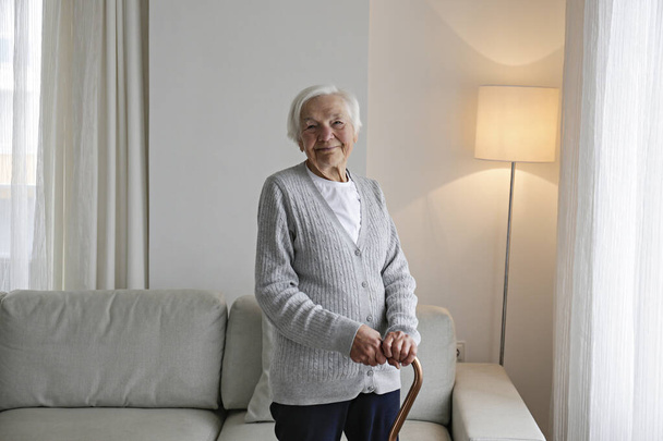 Senior woman 86 years of age standing by the window with walking cane in her apartment. Portrait of elderly lady in grey cardigan alone at home. Background, copy space, close up. - Fotoğraf, Görsel