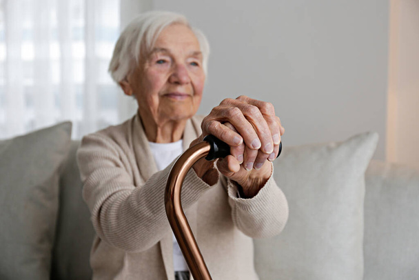 Portrait of a senior female sitting on the couch at home, leaning on a cane. Elderly woman holding the wooden handle of a metal walking cane. Close up, copy space for text, background. - Photo, image