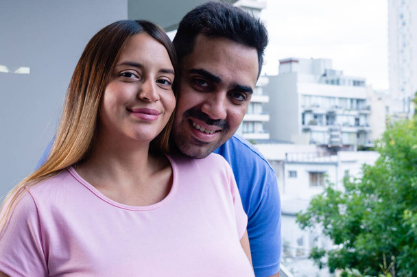 Porträt eines schwangeren heterosexuellen Paares, zu Hause auf dem Balkon lächelnd und in die Kamera blickend, junge kaukasische Männer und Frauen zusammen zu Hause. - Foto, Bild