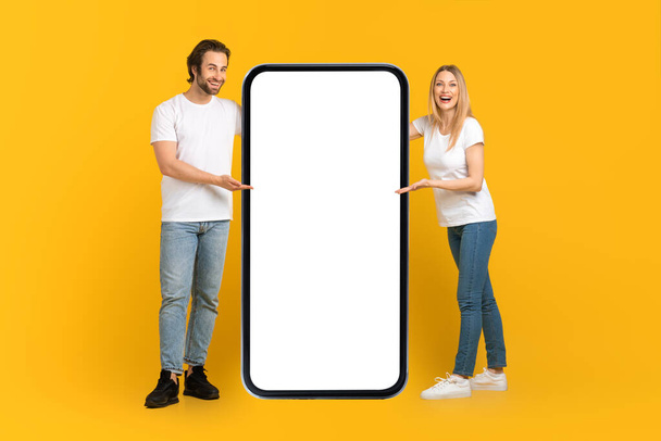 Happy young caucasian male and female in white t-shirts presentation huge phone with empty screen - Photo, Image