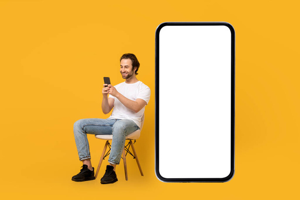 Smiling young european male in white t-shirt sits on chair, typing on phone near huge smartphone - Foto, imagen