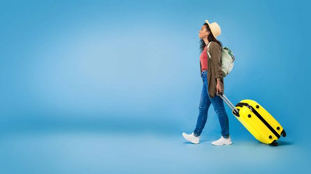 Feliz joven mujer negra caminando con maleta, viajando al extranjero, teniendo vacaciones de verano sobre fondo azul, espacio para copiar - Foto, imagen