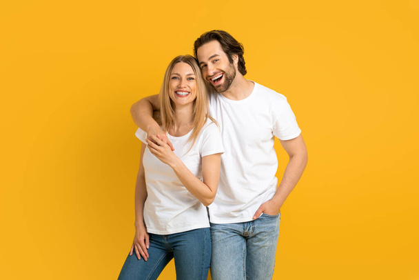 Boldog fiatal európai férfi és nő fehér pólóban ölelés, szórakozás, elszigetelt sárga háttér - Fotó, kép