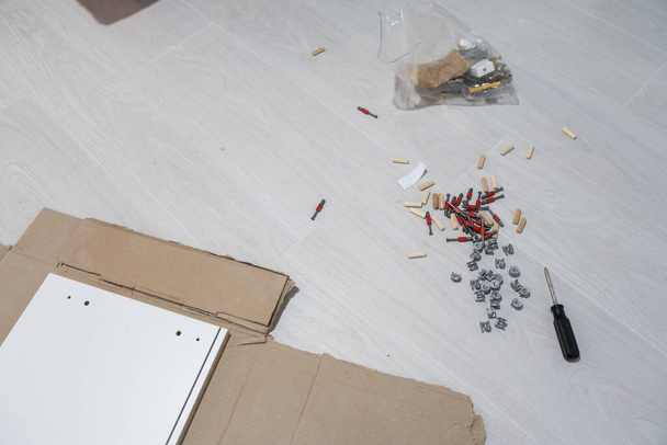 Parts of unfinished furniture, metal screws and tools lying on the floor with instruction manual for furniture assembly in the background. Moving to new home. - Fotografie, Obrázek
