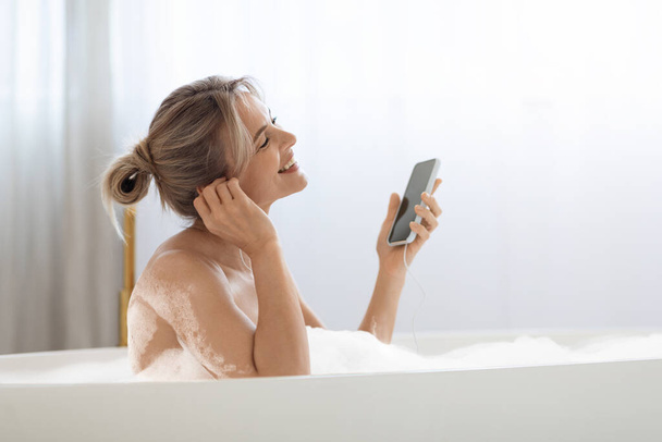 Mujer rubia positiva tomando un baño espumoso y escuchando música - Foto, imagen