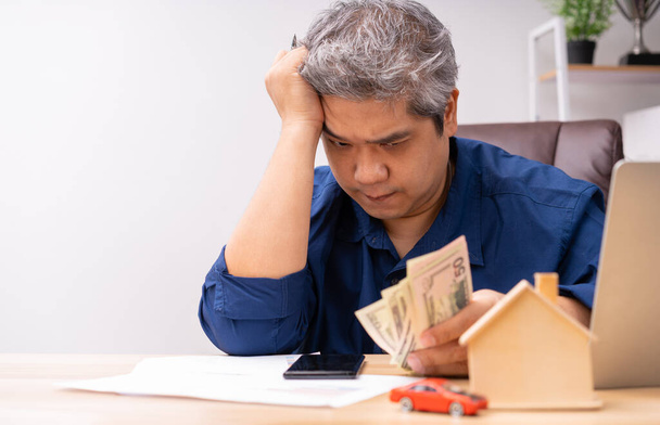Unhappy Asian man holding a banknote and Making an account of income and expenses to pay the home loan and car loan, Concept loan payment and home insurance, economic problems, and inflation - Photo, Image