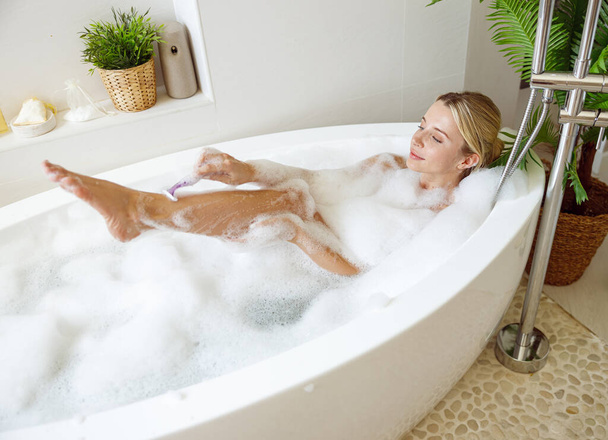 Beautiful woman shaving leg with razor in bathtub with foam. Relaxation and taking care. - Photo, Image