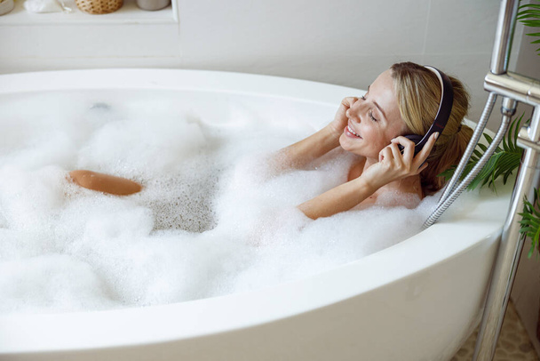 Cheerful young woman in headphones smiling, listening to music in bath with bubbles. - Foto, afbeelding
