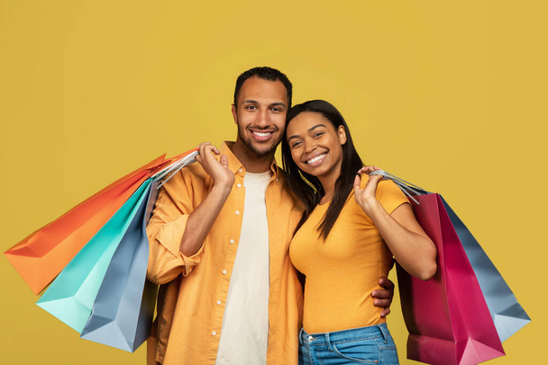 Glückliches junges schwarzes Paar mit Einkaufstüten, die lächelnd in die Kamera auf gelbem Studiohintergrund schauen - Foto, Bild