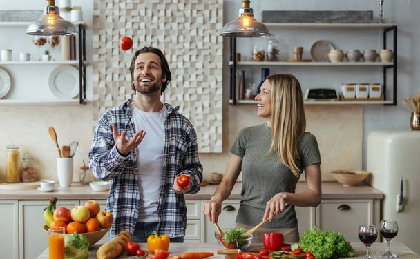 Glad caucasian millennial male with stubble juggles tomatoes, blonde lady prepares salad - Foto, imagen