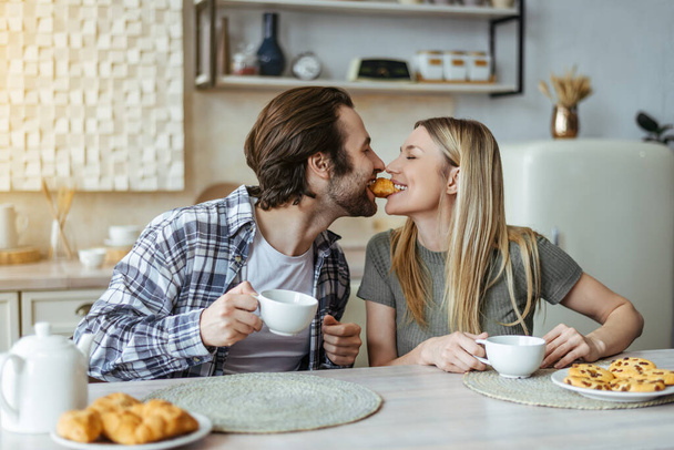 Glad european millennial couple eating cookies and drinking coffee, have breakfast in light kitchen - Фото, зображення