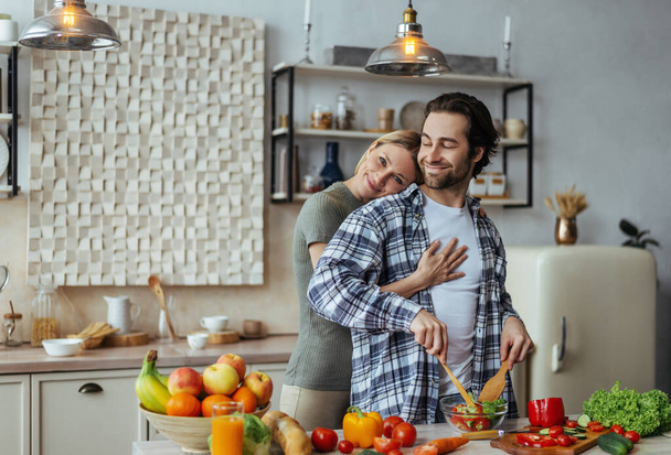 Sonriente caucásico millennial esposa abrazando hombre con rastrojo, chico preparando ensalada en mesa - Foto, imagen
