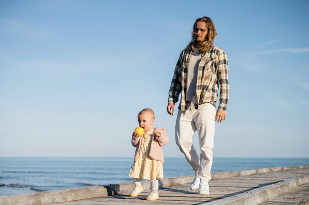 Young man walking near baby daughter with apple on pier on beach in Italy  - Foto, Imagen