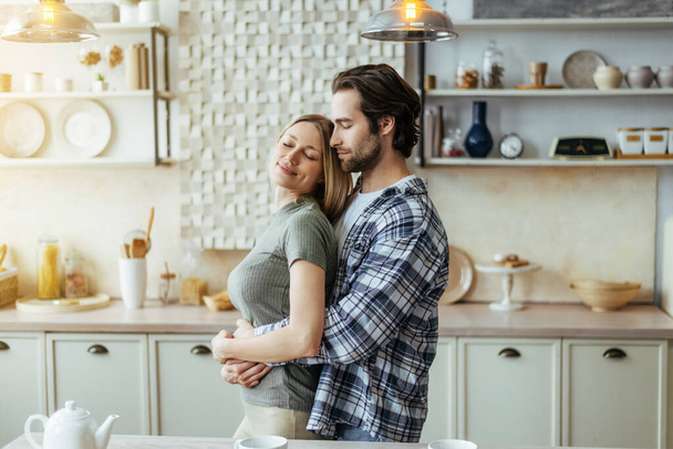 Vrolijk gelukkig Kaukasische millennial hasband met stoppelknuffels blonde vrouw, genieten van zacht moment in lichte keuken - Foto, afbeelding