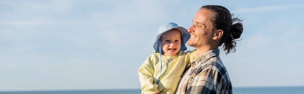 Papa en chemise tenant enfant en bas âge dans un chapeau panama avec la mer à l'arrière-plan, bannière  - Photo, image