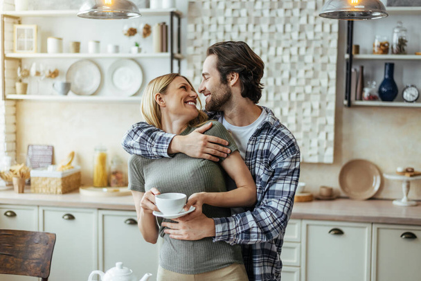 Smiling caucasian millennial male with stubble hugs blonde female, drink coffee, enjoy tender moment - Photo, Image