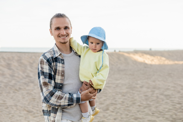 Positive man in shirt holding daughter and looking at camera on beach  - Zdjęcie, obraz