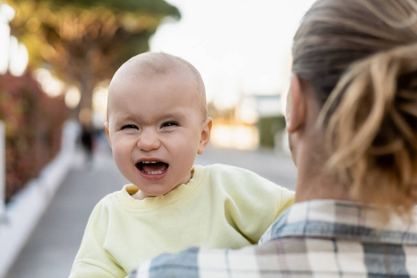 Menina bebê com boca aberta olhando para a câmera perto do pai ao ar livre  - Foto, Imagem