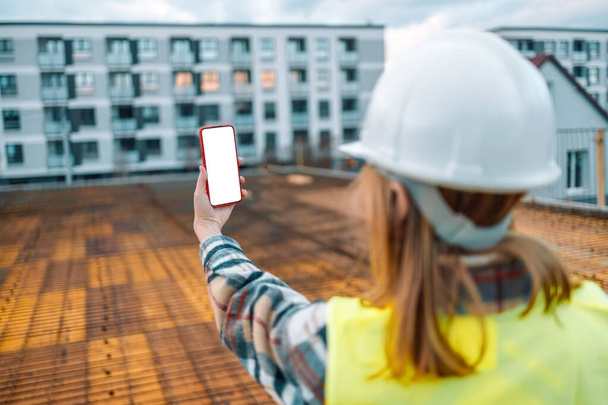 30s female architect with smartphone with white screen taking a photo at the construction site checking the progress of the construction work - Foto, Imagen