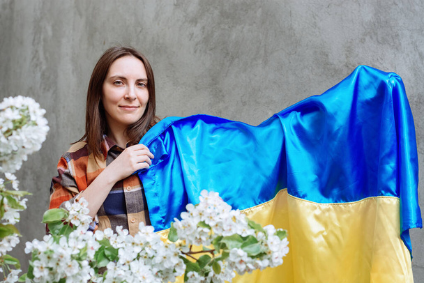 Schöne Mädchen hält in ihrer Hand die blau-gelbe Flagge der Ukraine auf dem Hintergrund einer grauen Betonmauer Stadt. Frau im karierten Hemd. Im Vordergrund verschwimmen Apfelblüten. Kopierraum. - Foto, Bild