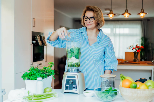 Middle aged woman with blender and green vegetables making detox shake or green smoothie at home. Healthy dieting, eating, cooking. Natural anti aging, weight loss program. Vegan, vegetarian diet - Photo, image