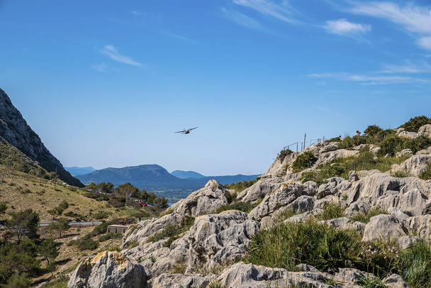 Airplane flying over rocky mountain. View of aircraft over landscape at island against blue sky. Scenic view of plants growing on cliff during summer. - Zdjęcie, obraz