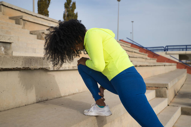 Femme afro-américaine avec des cheveux afro et des vêtements de sport, avec sweat-shirt jaune fluorescent et leggings, faire des étirements de jambe sur les escaliers. Concept fitness, sport, rue, urbain. - Photo, image