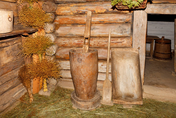 Antique wooden kitchen utensils - a mortar, a shovel for an oven and a trough in the hallway of an old rural log hut near tied bouquets of flax. - Fotó, kép