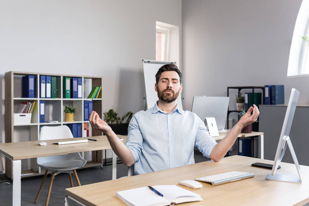 Young handsome man, office worker, sitting at a computer desk, stretching, tired, taking a break, doing yoga exercises, resting - Фото, изображение