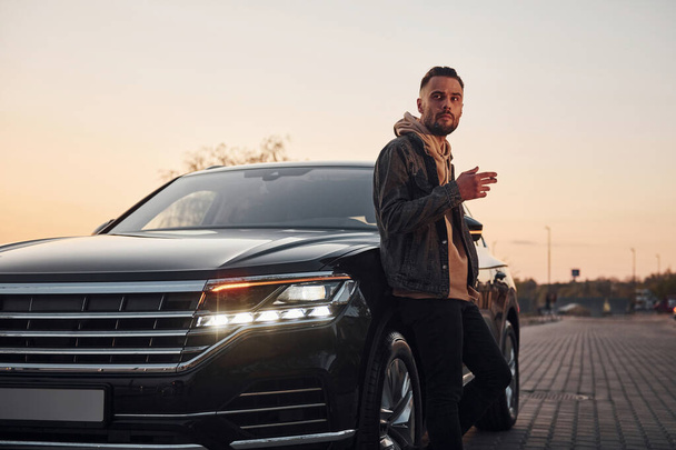 Handsome unshaved man in fashionable clothes standing near his black car and smoking. - Fotoğraf, Görsel
