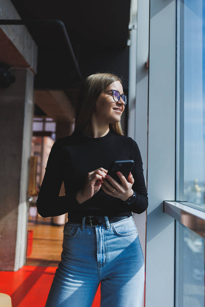 Mulher europeia alegre em óculos verificando e-mail de mídia social usando smartphone e conexão wi-fi, menina positiva compartilhar post do blog pessoal enquanto desfruta de aplicativo de telefone móvel - Foto, Imagem