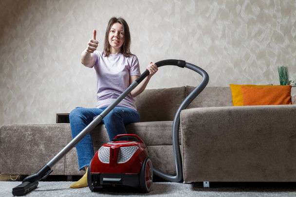 house cleaning concept. the vacuum cleaner is on the carpet next to the sofa. modern interior. the girl smiles and sits on the sofa. - Photo, Image