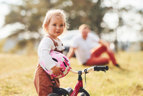 Father in white shirt sitting on the ground and his daughter standing in front of camera with her bike. - Photo, Image