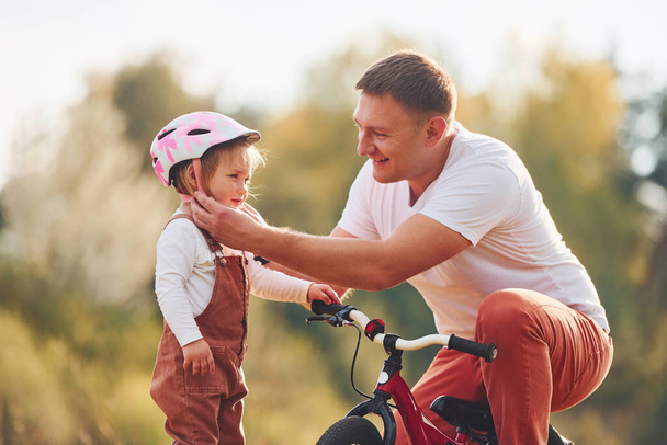 Beautiful sunlight. Father in white shirt teaching daughter how to ride bicycle outdoors. - Photo, Image