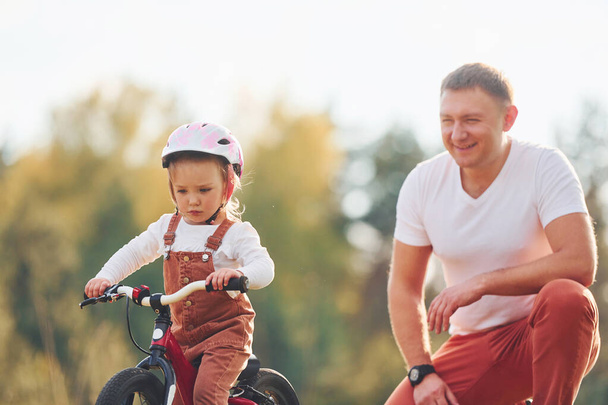 Beautiful sunlight. Father in white shirt teaching daughter how to ride bicycle outdoors. - Photo, Image