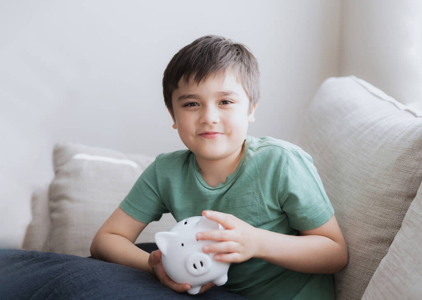 Happy boy holding piggy bank with smiling face. Indoor portrait of a cheerful child showing money saving box.School kid Learning financial responsibility and planning about saving for future concept - Photo, image