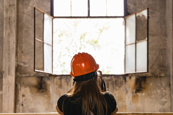 Woman in orange helmet on head standing in front of open window of old boiler building  - Foto, Bild