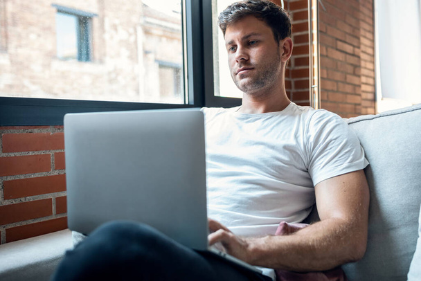 Tiro de homem inteligente trabalhando com seu laptop enquanto sentado em um sofá em casa - Foto, Imagem