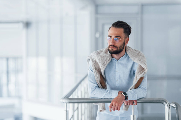 Front view of young handsome man in formal clothes that leaning on silver colored railings indoors in the office at daytime. - Фото, зображення