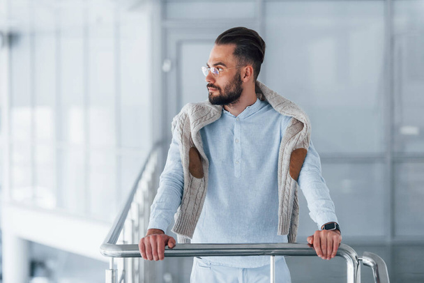 Front view of young handsome man in formal clothes that leaning on silver colored railings indoors in the office at daytime. - Фото, зображення