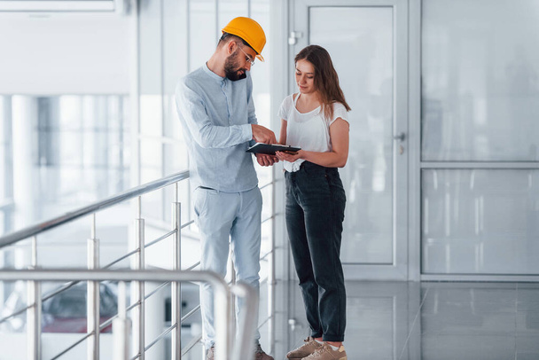 Talking with young girl in casual clothes. Engineer in white clothes and orange protective hard hat standing and working indoors. - Photo, Image